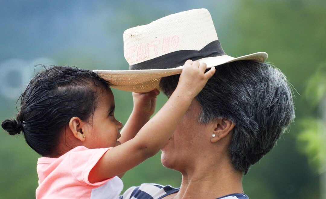 Frau mit Kind auf dem Arm. Das Kind hält einen Strohhut über den Kopf der Frau. Kommunikation mit dem inneren Kind.